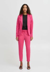 Rosa taillierte Blazerjacke und passende Jogginghose mit Kordelzug, kombiniert mit einem schwarzen Oberteil. Glatte Textur, maßgeschneidertes Design und minimale Hardware.