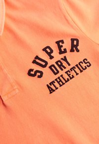 Pomarańczowa koszulka polo z teksturowanej tkaniny. Ma czarny haftowany napis "SUPER DRY ATHLETICS" z przodu. Klasyczny design kołnierza.