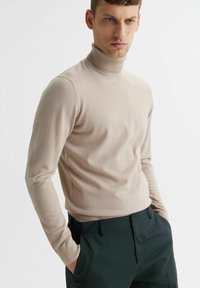 Beżowy sweter z golfem o dopasowanym kroju, z żebrowanym dekoltem i długimi rękawami, połączony z ciemnozielonymi spodniami. Miękka tekstura, minimalne detale.