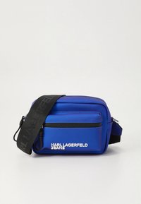 Modrá nylonová kabelka přes rameno s jednou hlavní přihrádkou a přední kapsou na zip. Má černý popruh s nápisem "KARL LAGERFELD JEANS".