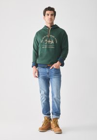 Sweat à capuche vert avec un motif d'ours, jeans et bottes beiges. Le sweat est équipé d'un cordon de serrage et d'une poche avant. Un look décontracté avec un style superposé.