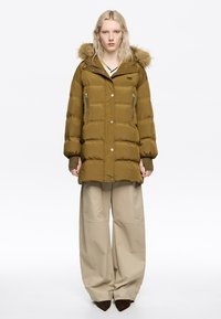 Chaqueta acolchada verde oliva con capucha forrada de piel, bolsillos con cremallera y botones a presión. Se lleva sobre pantalones beige anchos. Textura suave.