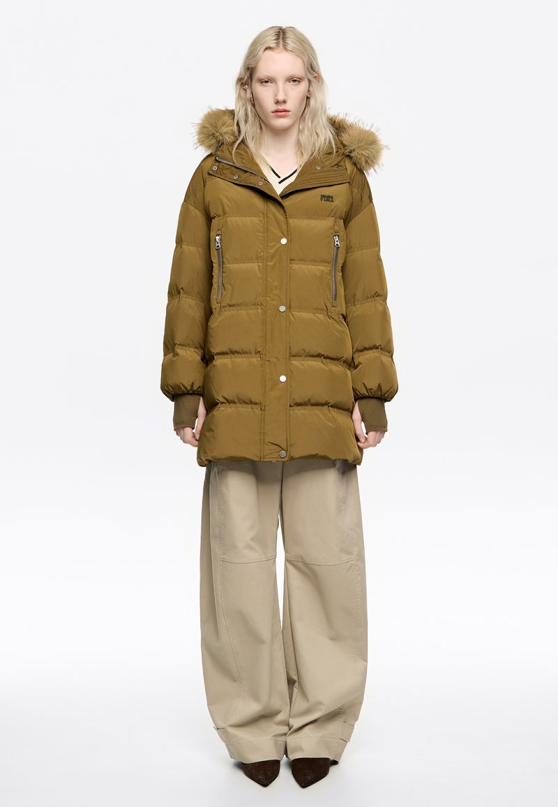 Chaqueta acolchada verde oliva con capucha forrada de piel, bolsillos con cremallera y botones a presión. Se lleva sobre pantalones beige anchos. Textura suave.