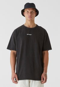 Black oversized t-shirt with "LOST YOUTH" emblem in white, short sleeves, round neckline, and relaxed fit. Worn with a black bucket hat.