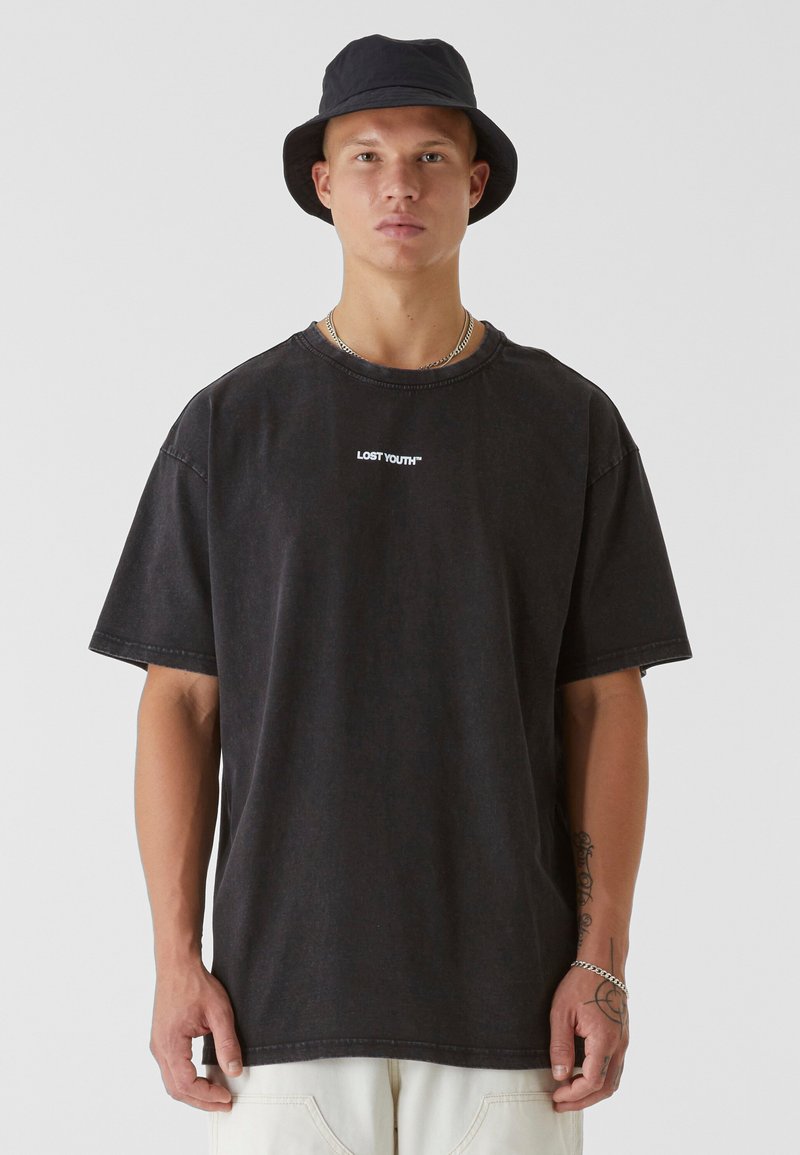 Black oversized t-shirt with "LOST YOUTH" emblem in white, short sleeves, round neckline, and relaxed fit. Worn with a black bucket hat.