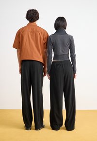 Deux individus de dos, portant une chemise orange à manches courtes et une veste noire courte, tous deux en pantalons noirs amples et des chaussures.