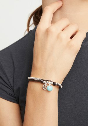 Bracelet en cuir orné de perles argentées, d'une perle turquoise et d'un pendentif symbole de la paix en argent. Porté à la main contre une chemise foncée.