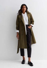 Trench coat verde oliva con bottoni doppi, maniche lunghe e vita con cintura. Abbinato a una camicia bianca e pantaloni scuri.