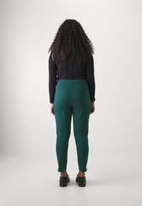Green tailored trousers with pockets, slim fit, and straight legs. Worn with a black long-sleeve top and black shoes.