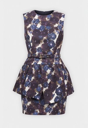 Vrouw met lang donker haar, die een mouwloze bloemenjurk met riem en zwarte hakken draagt, staat met één hand op haar heup.