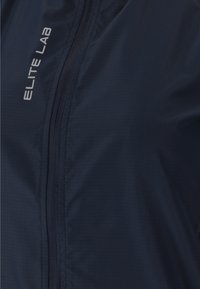 Marineblå vindjakke med struktureret finish, der har en frontlynlås, "ELITE LAB" logo i grå og en tætsiddende design. Velegnet til udendørs brug.