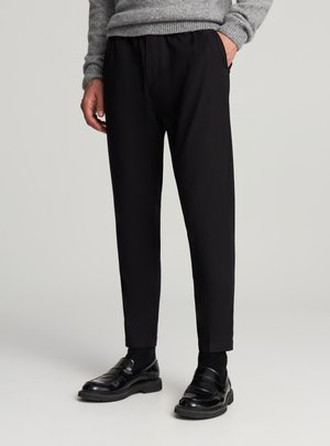 Pantaloni neri con vestibilità su misura, vita elasticizzata, tasche laterali e una texture liscia, abbinati a mocassini neri lucidi.