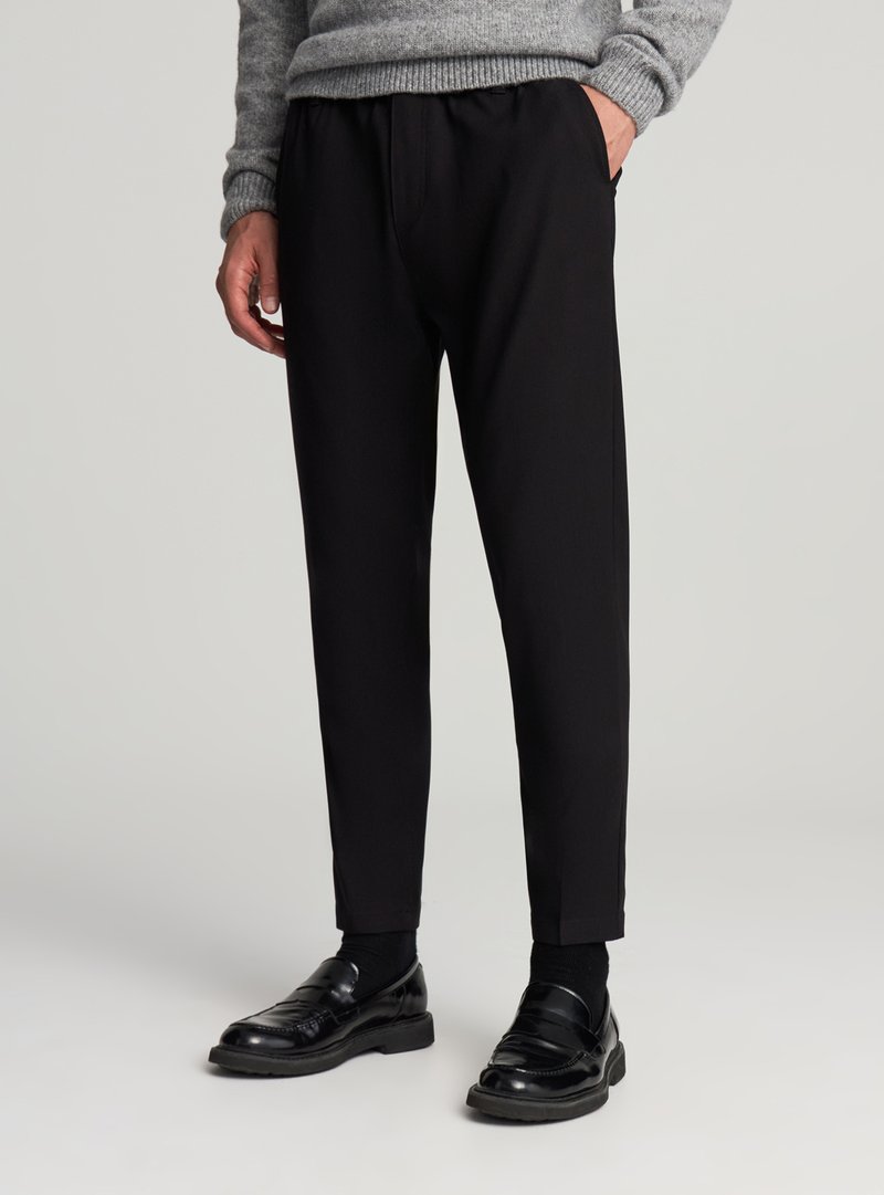 Pantaloni neri con vestibilità su misura, vita elasticizzata, tasche laterali e una texture liscia, abbinati a mocassini neri lucidi.