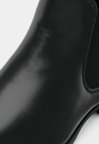 Bottine en cuir noir à fini brillant, avec des panneaux latéraux élastiques et un bout arrondi, présentant une texture lisse et un design minimaliste.