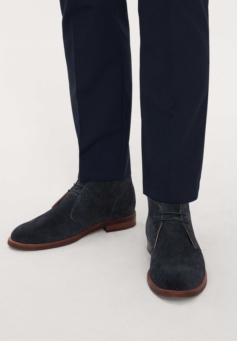 Bottines en daim bleu foncé avec semelles marron portées avec un pantalon bleu marine foncé sur un sol gris clair.