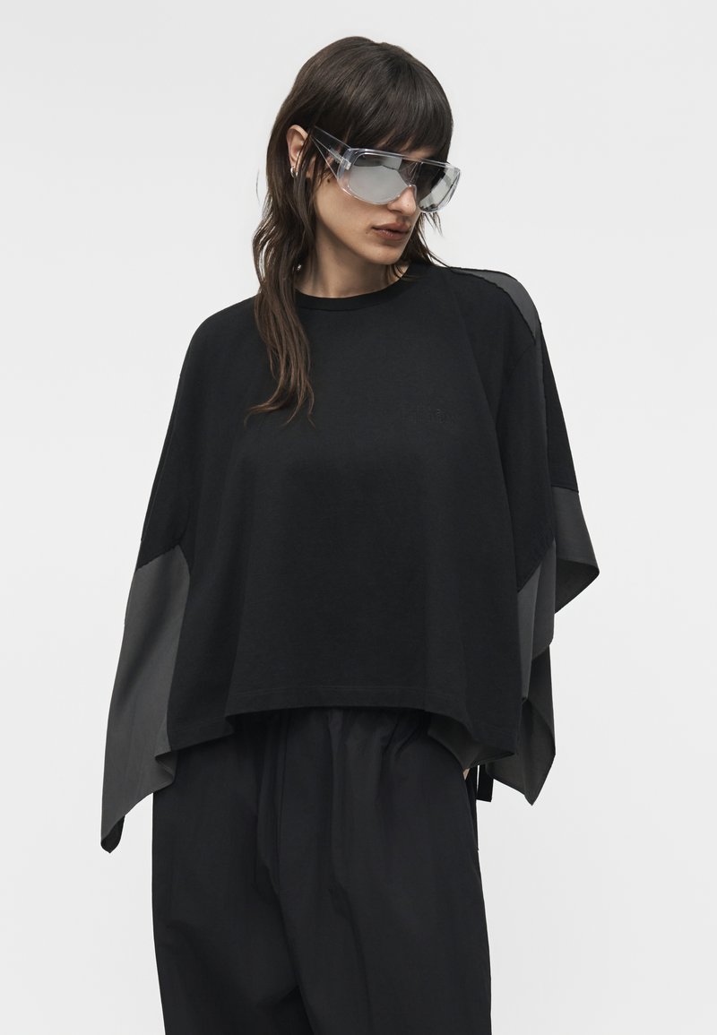 Femme portant un haut noir ample à manches larges et de grandes lunettes de soleil transparentes, regardant légèrement vers le bas et sur le côté.