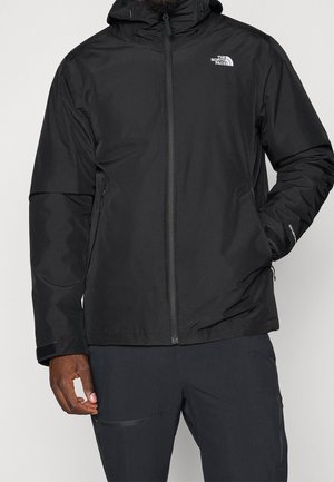 Mann trägt schwarze North Face Kapuzenjacke mit Reißverschluss vorne und seitlichen Reißverschlusstaschen, eine Hand in der Tasche, steht vor einfachem Hintergrund.