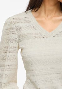 Light beige long-sleeve top featuring a V-neck, textured ribbed material, and horizontal stripes of varying thickness and openness.