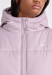 Gai lavandas krāsas pūšamā jaka ar rāvējslēdzēja aizdari, augstu apkakli un baltu Nike logotipu uz krūtīm. Mīksta, gluda faktūra un kvadrātveida dizains.