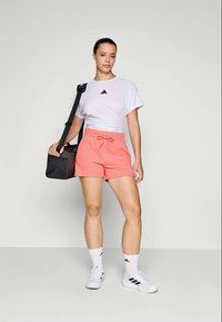 T-shirt branca da Adidas com logótipo preto, calções corais e mala de desporto preta. Sapatos brancos de desporto e meias até ao tornozelo com logótipo da Adidas. Design leve.