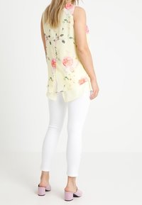 Světlounce žlutá tílková halenka s květinovým potiskem; rozparky na zádech. Kombinováno s bílými přiléhavými kalhotami a růžovými mules s blokovým podpatkem.
