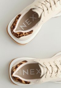 Beige tenisky s texturovaným textilním svrškem, leopardími akcenty na patě a bílou podešví s průhlednou gumovou základnou.