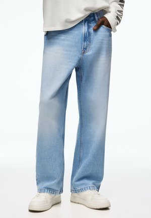 Person trägt hellblaue Jeans mit weitem Bein, weiße Sneakers und ein weißes Langarmshirt, eine Hand in der Tasche.