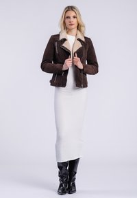 Blonde vrouw in een bruine suède jas over een witte jurk, zelfverzekerd staand in zwarte kniehoge laarzen tegen een effen achtergrond.