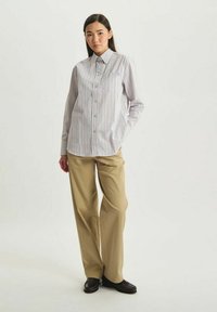Chemise à boutons en blanc avec des rayures verticales pastel ; elle présente un petit logo et une seule poche poitrine. Associée à un pantalon beige.