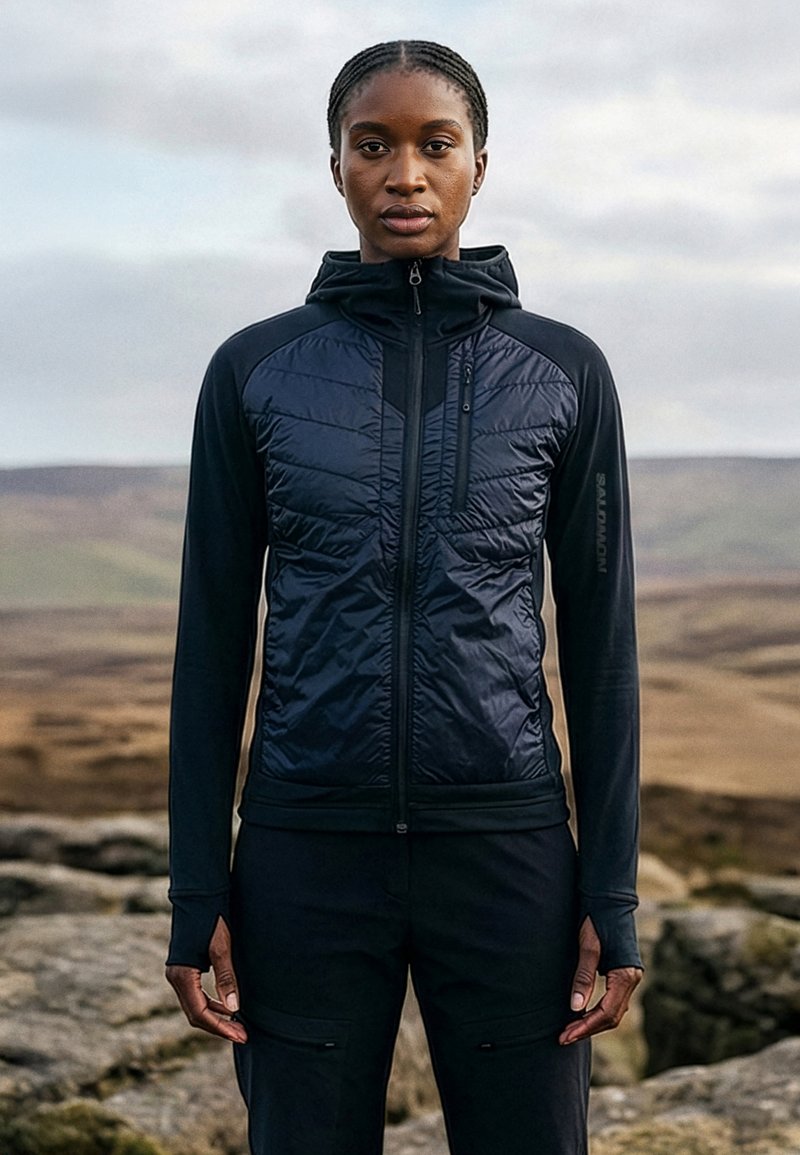 Schwarze Frau mit geflochtenem Haar, die eine schwarze gesteppte Jacke und Hose trägt, steht im Freien auf felsigem Gelände unter einem bewölkten Himmel.