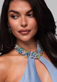 Blå halterneck-top som har ett flerfärgat stenhalsband med blå, gula och rosa detaljer, mot en slät hud och mörkt hår.