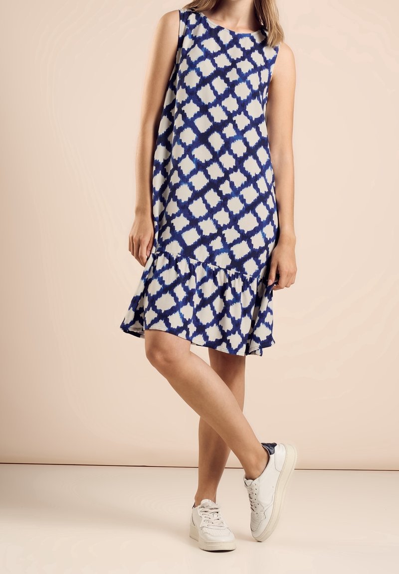 Robe sans manches à motifs bleus et blancs avec un ourlet à volants, associée à des baskets blanches, sur un fond clair.