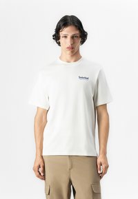 White cotton t-shirt with a crew neck and short sleeves. Features a blue Timberland logo on the left chest. Smooth texture, regular fit.