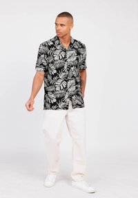 Camisa negra de manga corta con un patrón de hojas blancas, combinada con pantalones color crema y zapatillas blancas. El fondo liso resalta el enfoque en la vestimenta.