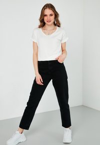 Hvid kortærmet V-hals t-shirt parret med sorte højtaljede jeans og hvide sneakers, placeret mod en ensfarvet baggrund.