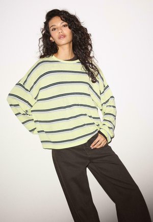 Donna con capelli ricci che indossa un maglione giallo a righe ampio e pantaloni larghi scuri, posando contro uno sfondo chiaro e semplice.