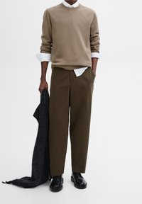 Beige gestrickter Pullover, darunter ein weißes Hemd mit Kragen, olivgrüne Hose, schwarze Schuhe und ein grauer Schal über dem Arm drapiert.