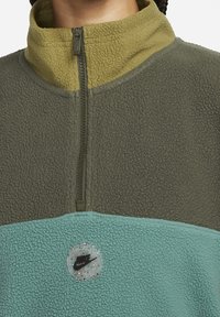 Fleece-polerad tröja i mörkgrön och mintblå med hög krage, dragkedjestängning och ett runt, flerfärgat Nike-logopatch i mitten.