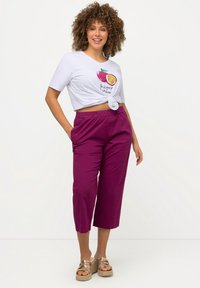 Witte cropped t-shirt met een fruitafbeelding en tekst, gecombineerd met paars broek tot halverwege de kuit. Model draagt tan platform sandalen.