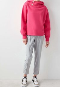 Sweatshirt rose avec un col montant et des boutons décoratifs, associé à un pantalon sur mesure gris clair et des baskets noir et blanc.