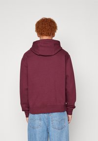Sudadera burdeos con una gran capucha en la parte posterior, puños y dobladillo acanalados. Tela suave y texturada. Combinada con jeans de un azul claro.