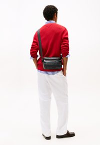 Borsa a tracolla nera con due compartimenti con zip, realizzata in pelle liscia, indossata sulla spalla, abbinata a un maglione rosso e pantaloni bianchi.