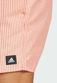 Pantaloni corti a righe rosa e crema con un tessuto testurizzato; presenta una patch nera del logo Adidas vicino all'orlo.