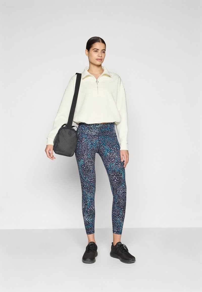 Helle cremefarbene Sweatjacke mit Reißverschluss, gemusterte blaue und lila Leggings und schwarze Sportschuhe. Das Modell hält eine kleine schwarze Tasche.