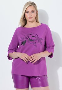 Paarse top met 3/4 mouwen en een zwart bloemmotief. Gepaard met glanzende paarse shorts. Glad textuur, ronde halslijn en een ontspannen pasvorm.