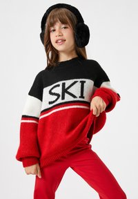 Roter und schwarzer Strickpullover mit dem Aufdruck "SKI" und weißen Streifenakzenten. Kombiniert mit roten Hosen trägt das Model schwarze Ohrenschützer.
