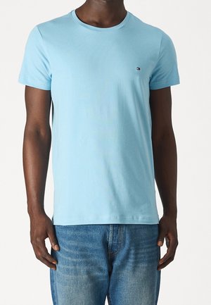 Homme portant un t-shirt bleu clair à manches courtes et col rond avec un petit logo sur la poitrine, associé à un jean denim bleu moyen.