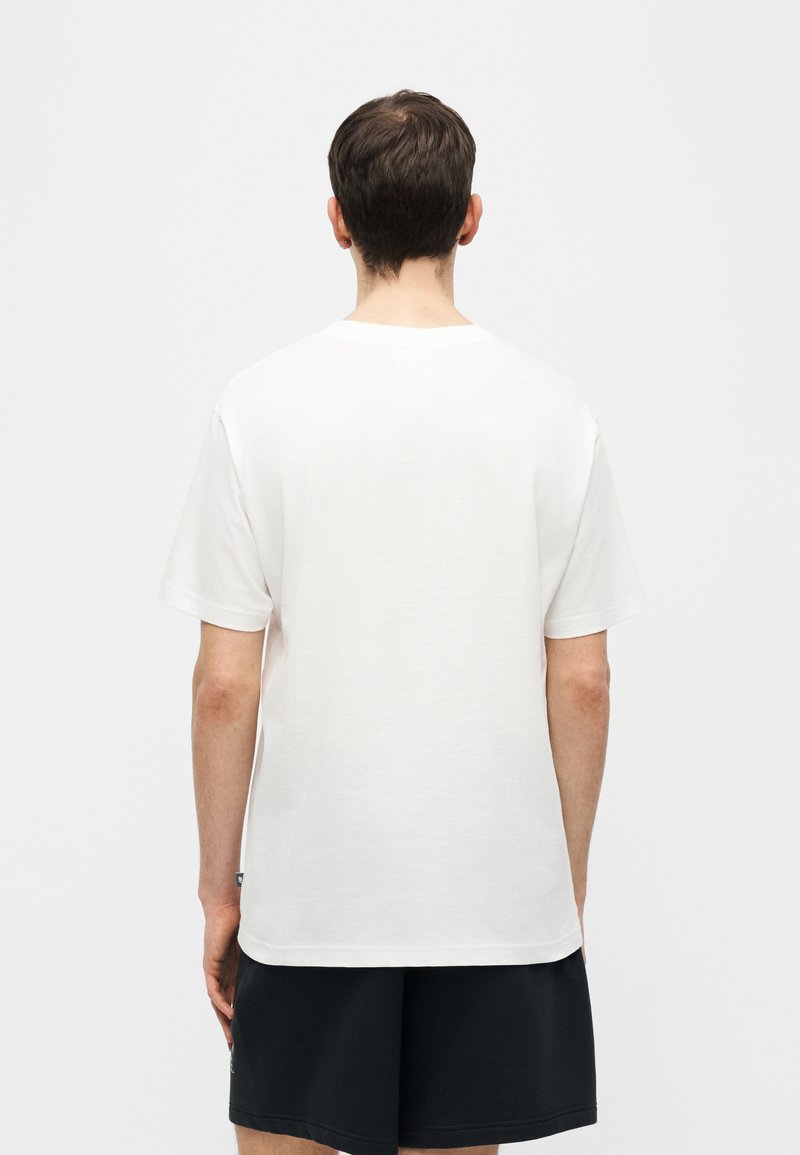 Personne portant un T-shirt blanc uni à manches courtes et un short noir, debout face à un fond blanc uni.