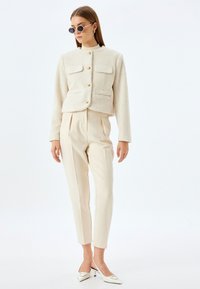 Veste crème texturée avec des boutons dorés et deux poches avant, associée à un pantalon beige ajusté et des mules à talons blancs.