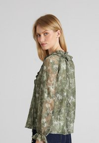 Durchsichtige grüne Bluse mit floralem Muster, Rüschenausschnitt und elastischen Bündchen. Leichter Stoff mit strukturierter Oberfläche und lockerer Passform.